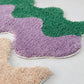 Three colorful bath mats in purple, green, and beige on a light gray surface.