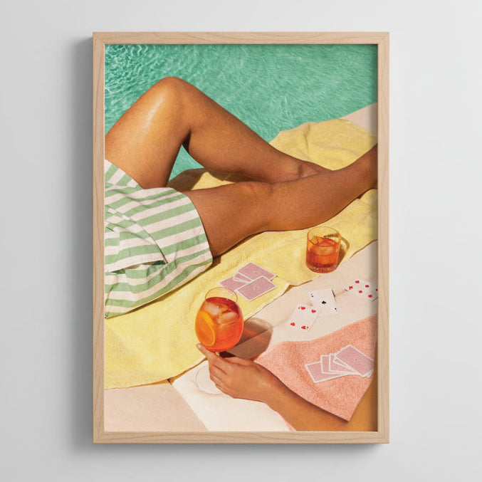 Person lounging by a pool with drinks and playing cards on a towel