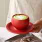 Red cup of green tea with latte art on a red saucer, held by a person.