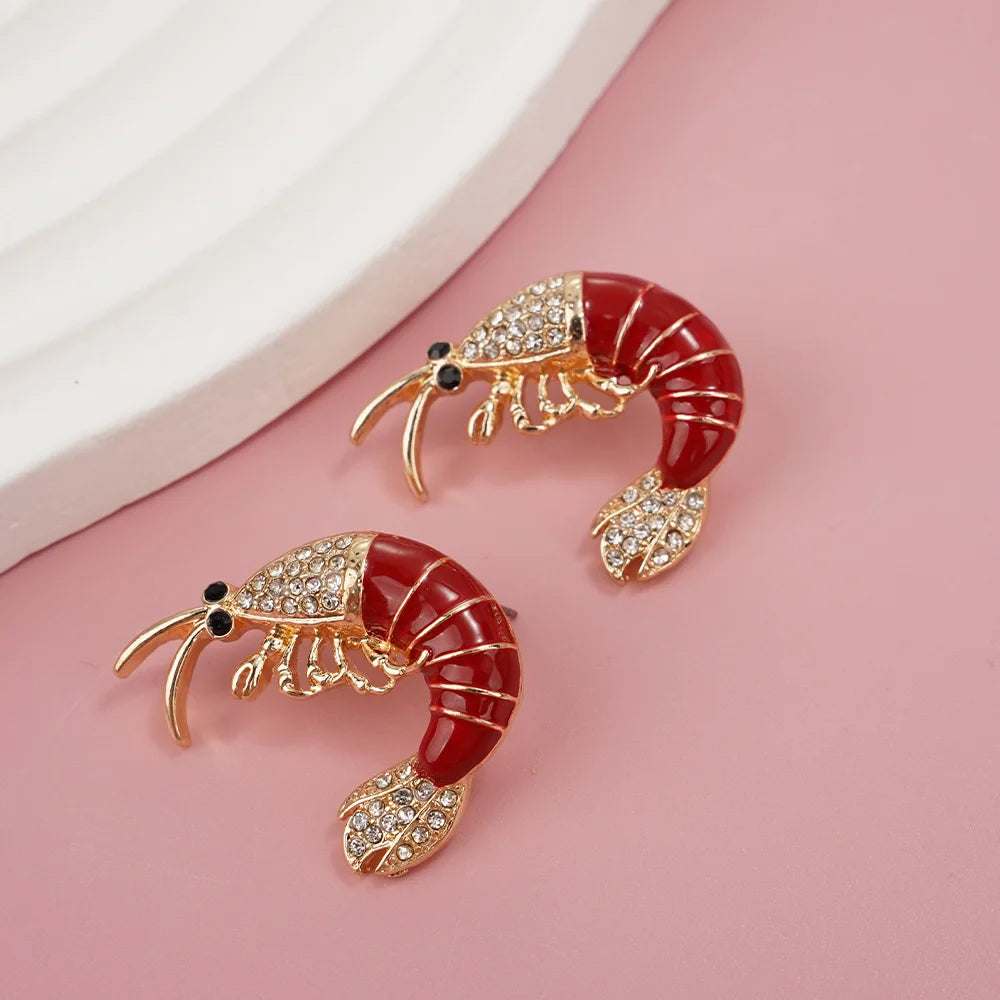 Two shrimp-shaped earrings with red bodies and gold accents on a pink background