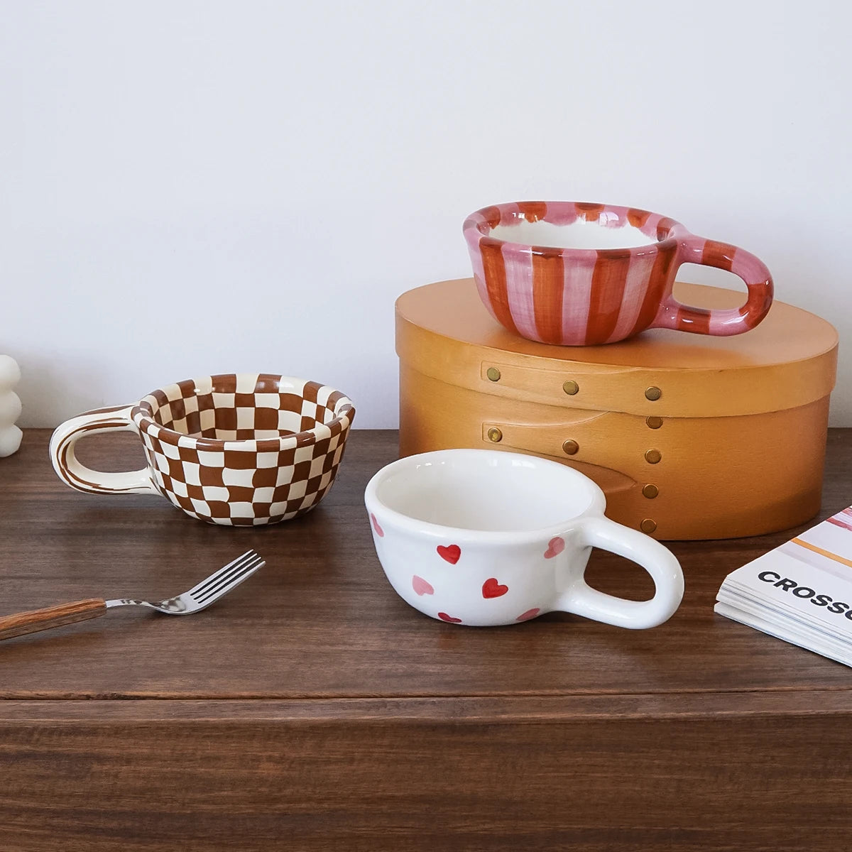 Set of ceramic mugs and bowls on a wooden surface with a fork and newspaper.
