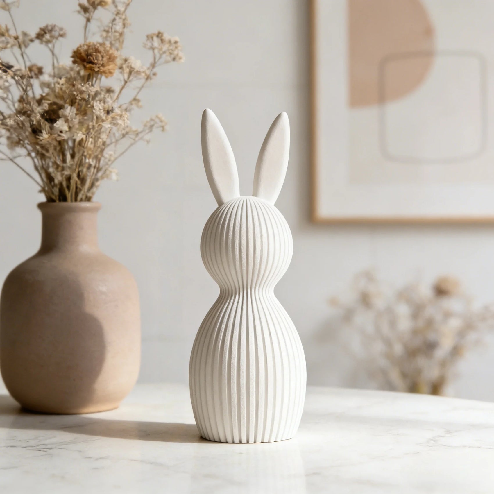 White rabbit-shaped vase on a marble surface with a beige vase and dried flowers in the background.