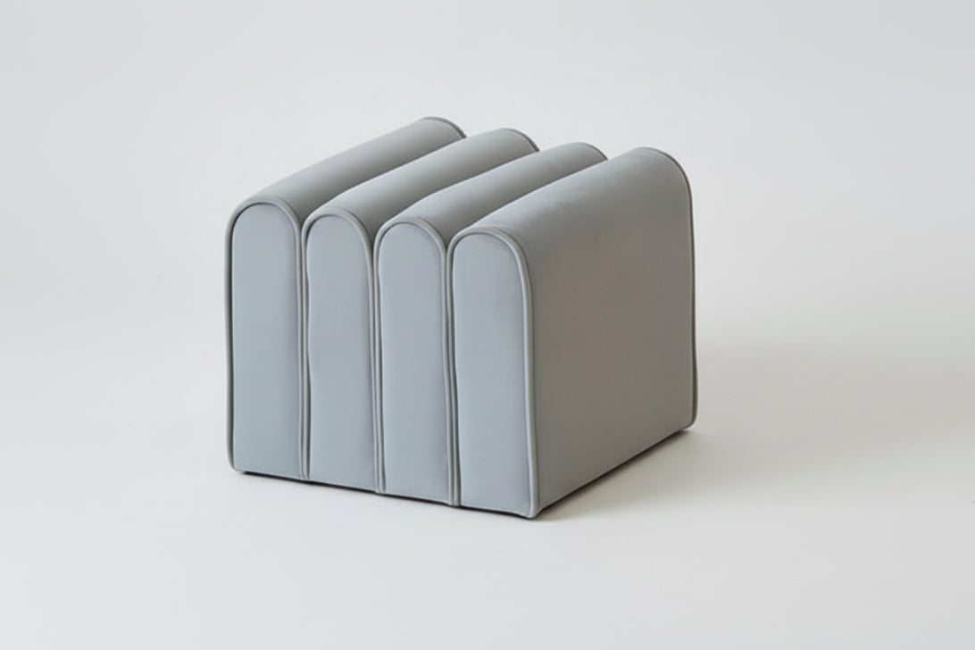 A modern gray and brown shoe change stool with a minimalist design, accompanied by a small lamp, placed next to a bookshelf with some books.