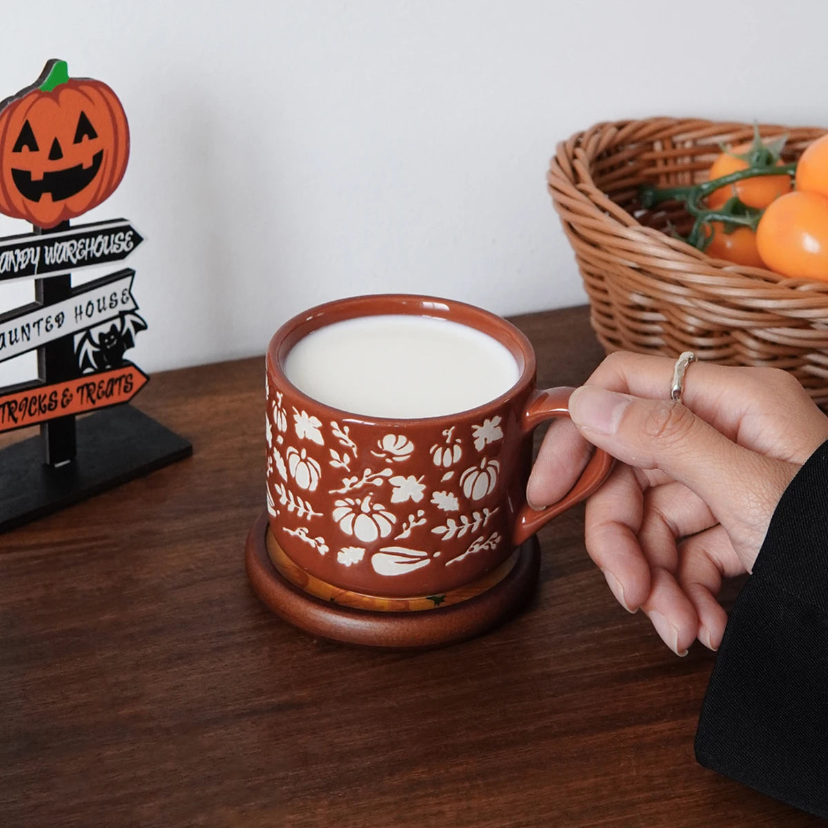 Person holding a decorative mug with pumpkins and floral patterns on a wooden table.
