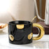 Black mug with gold handle and decorative elements on a reflective surface.