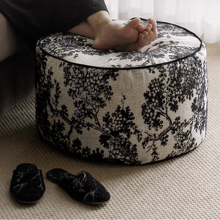Person sitting on a patterned ottoman surrounded by black slippers.