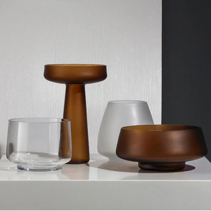 Three decorative vases in brown, clear, and white on a white surface with a gray wall background.
