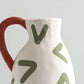 Ceramic pitcher with red handle and green abstract designs on a light gray background