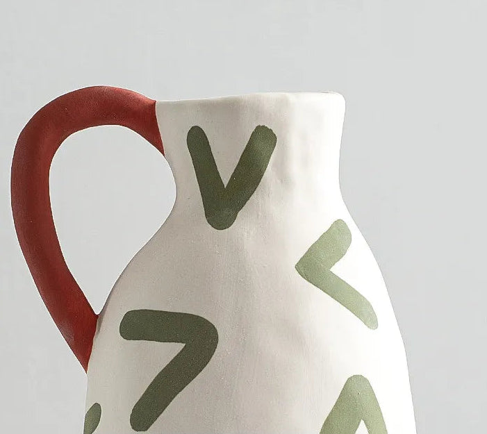 Ceramic pitcher with red handle and green abstract designs on a light gray background