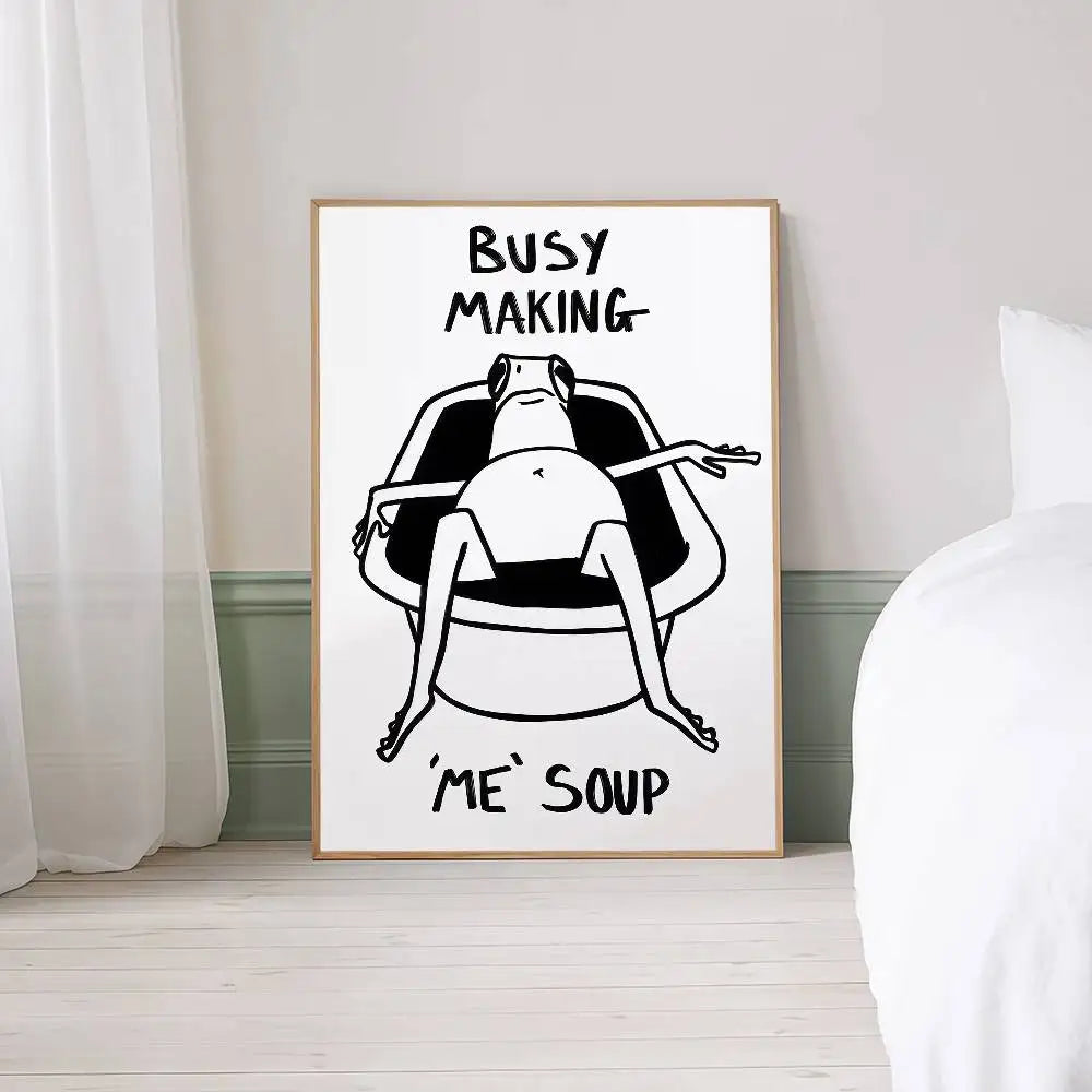 Framed artwork with a black and white illustration of a person in a bathtub and text 'BUSY MAKING ME SOUP' on a white wall.