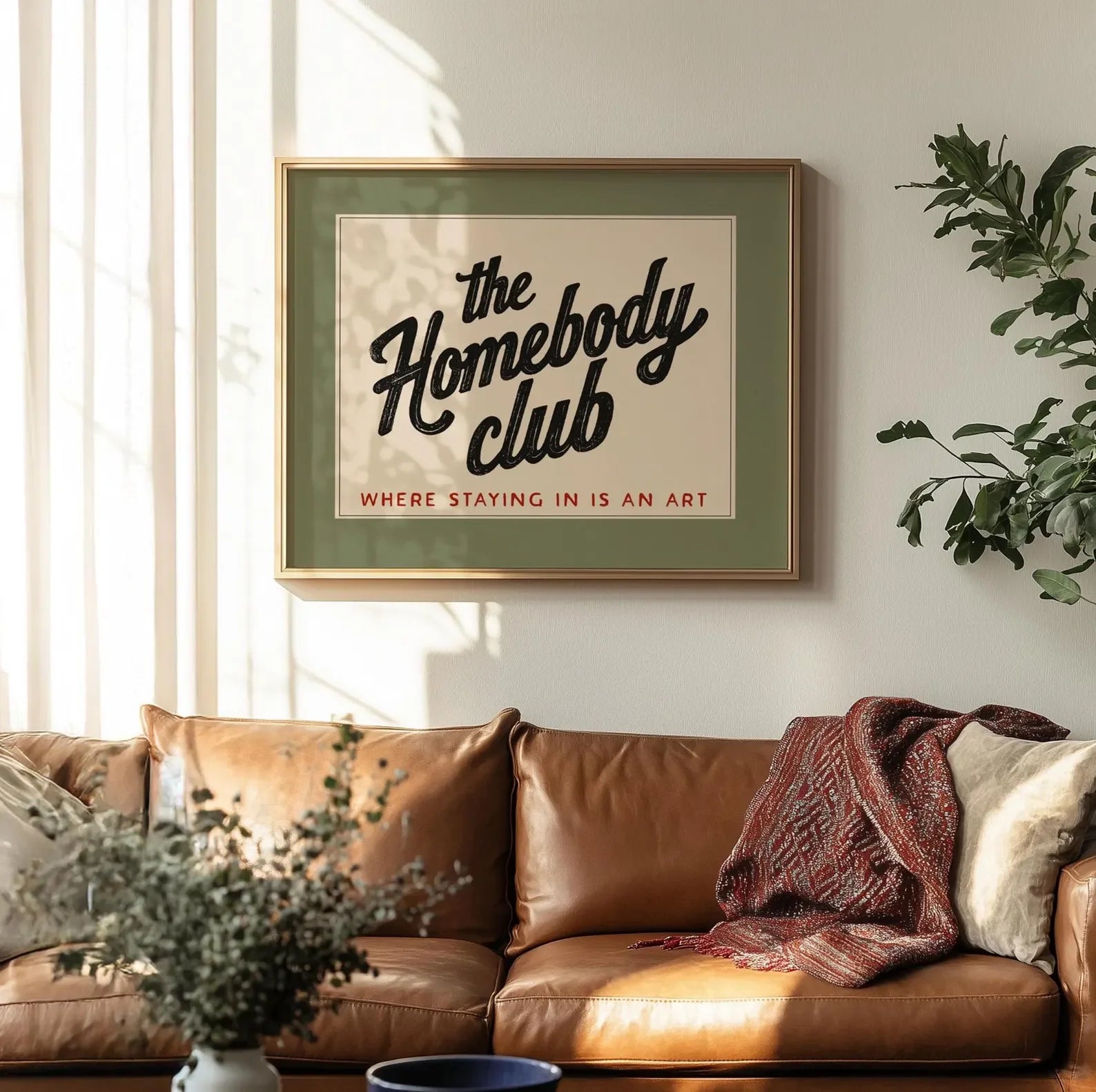 Framed artwork titled 'the Homebody Club' above a brown leather sofa with decorative pillows and a plant.