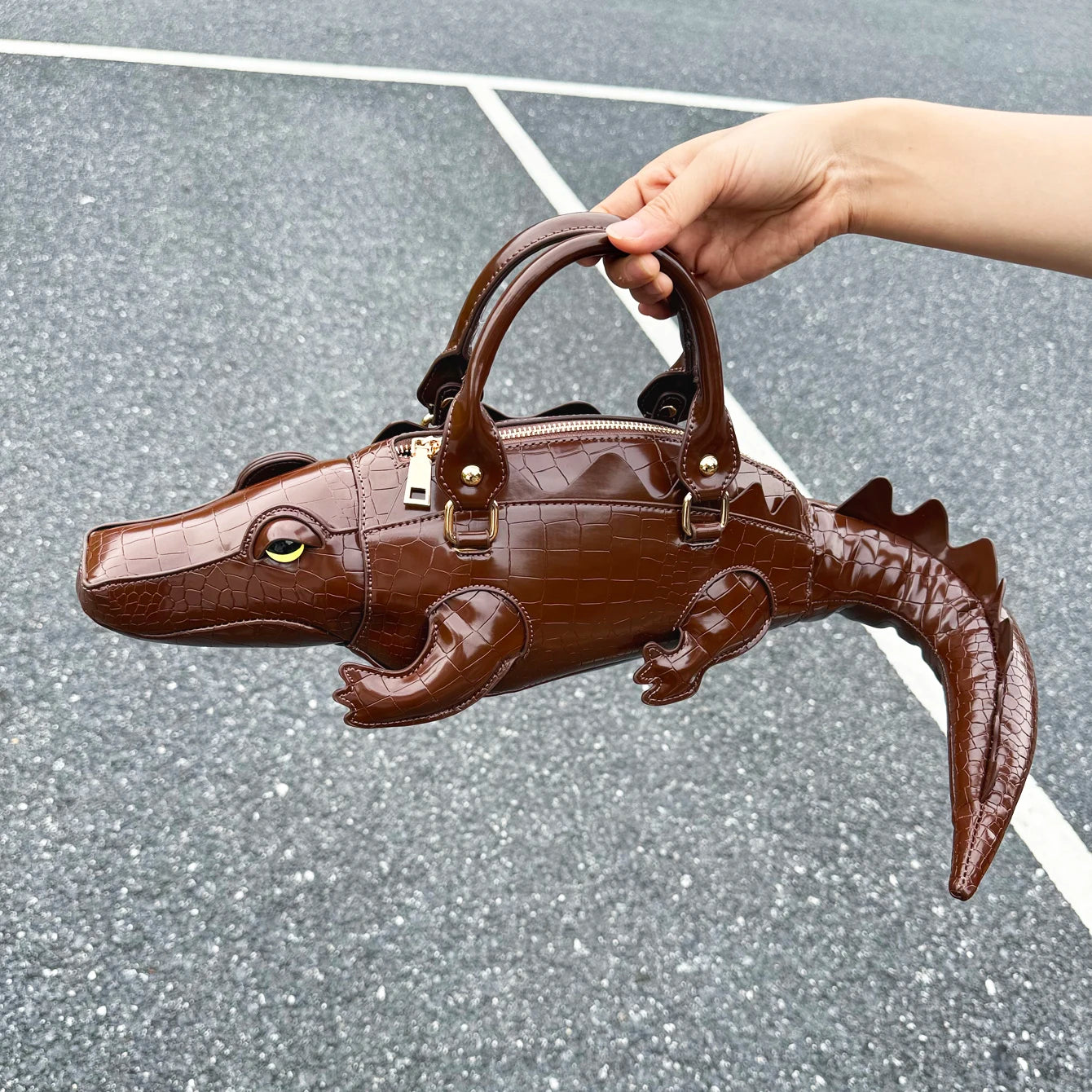 Brown alligator-shaped handbag held by a hand on a gray surface.
