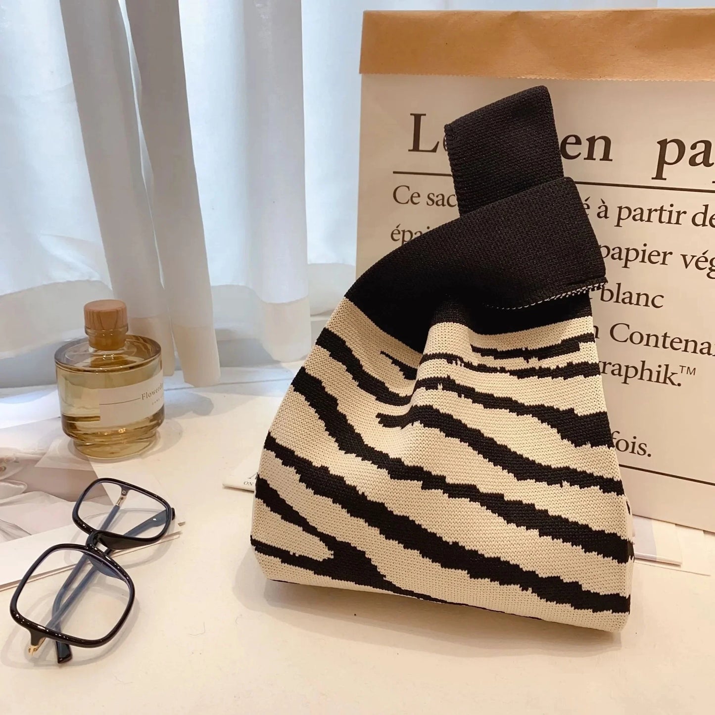 Black and white striped bag on a surface with glasses, perfume bottle, and paper in the background.