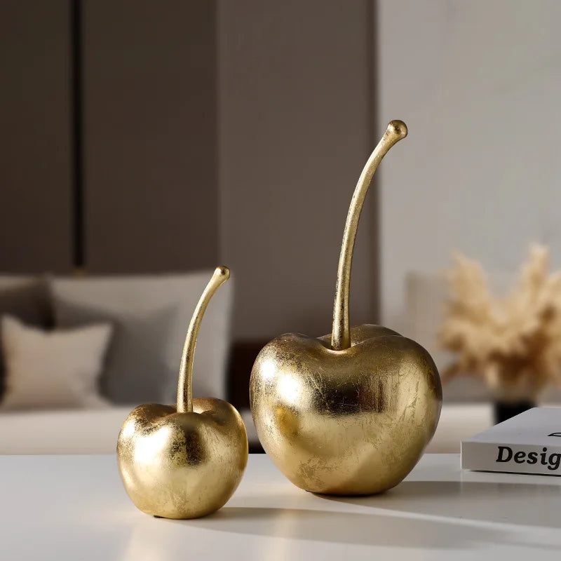 Two gold cherry decorations on a surface with a blurred indoor background