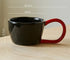 Black mug with red handle on a wooden surface, showing measurements and weight capacity.