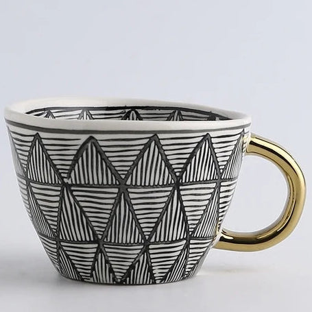 Ceramic mug with geometric black and white pattern and gold handle on a light gray background
