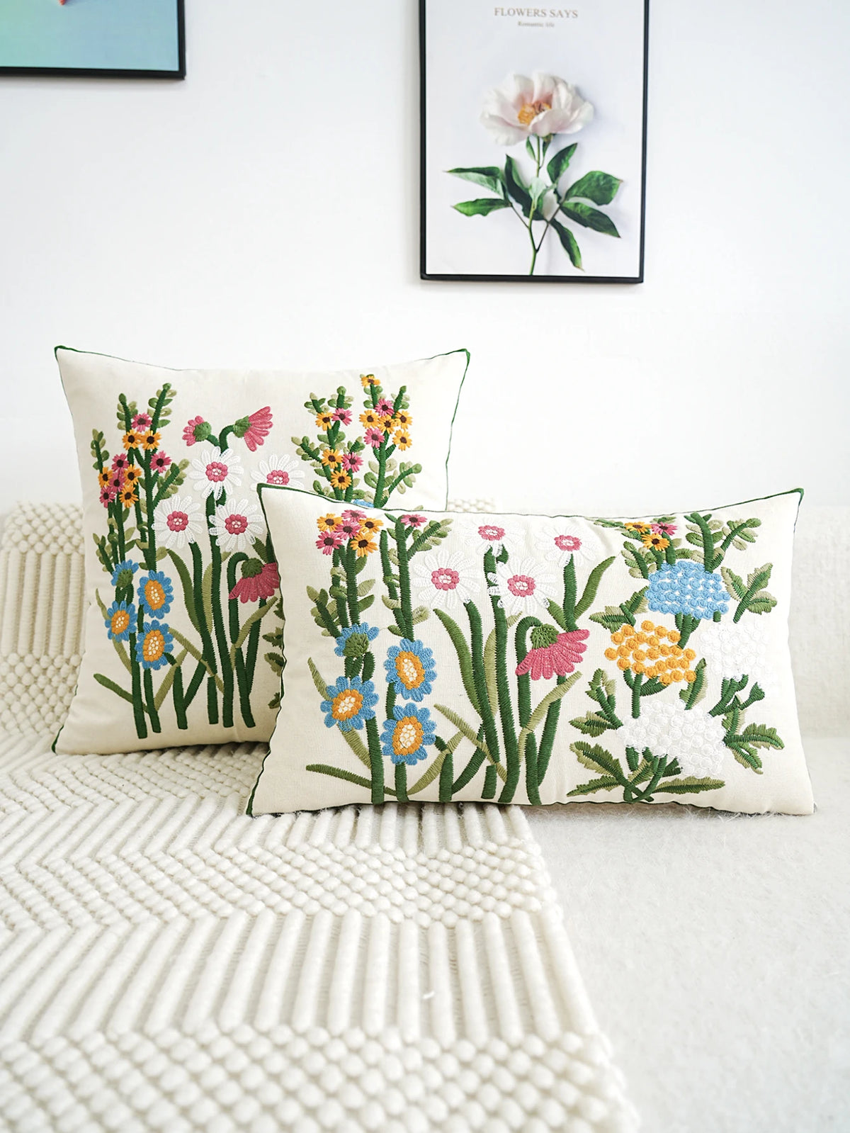 Two floral-patterned pillows on a bed with a white wall and framed artwork in the background.
