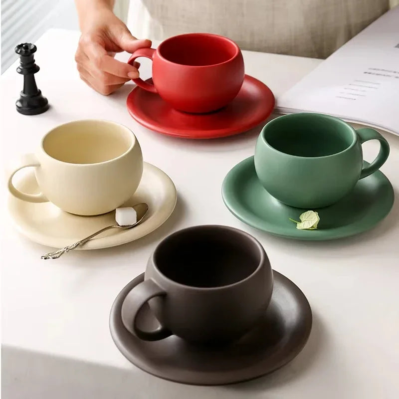 Four ceramic cups and saucers in red, green, beige, and black on a white surface.