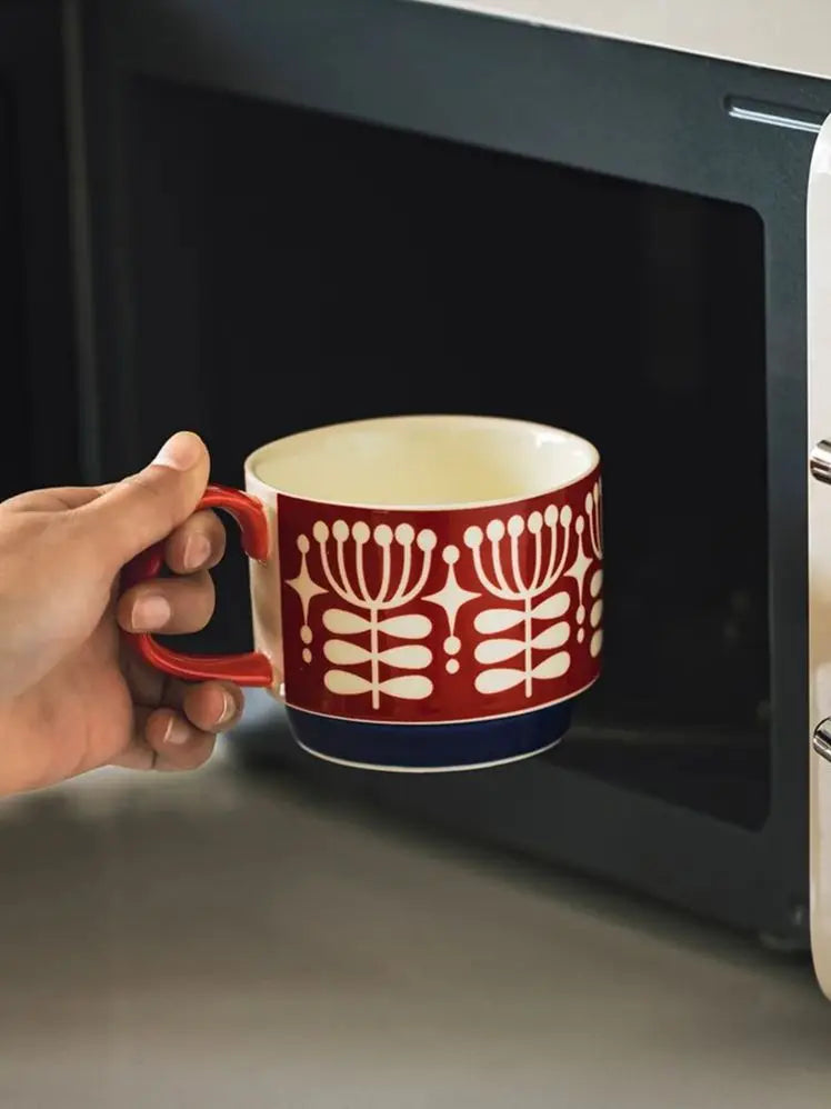 Hand holding a red and white mug with floral design in front of an open microwave.