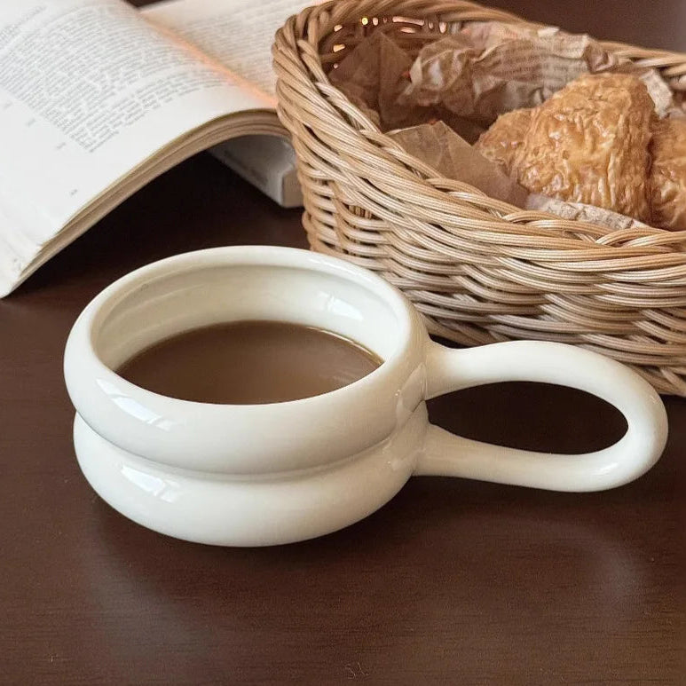White ceramic mug with handle on a wooden surface, containing a dark liquid, next to an open book and a woven basket with bread.