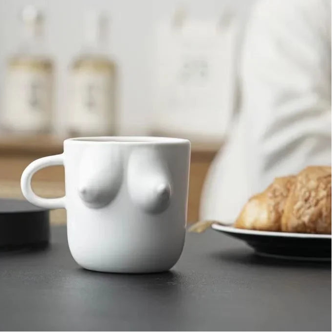 White mug shaped like a breast on a kitchen counter with a plate of cookies and coffee maker in the background.