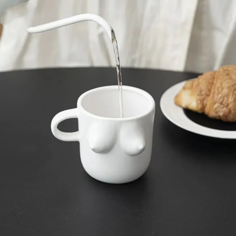 White mug shaped like a face with a nose, pouring liquid onto a black surface with a croissant on a plate in the background.