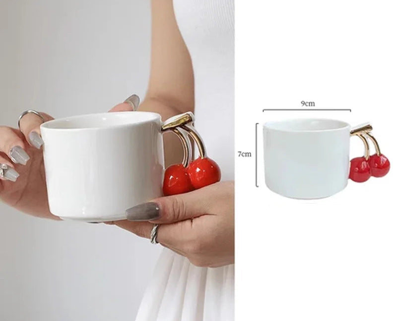White mug with cherry and gold handle held by a hand, with measurement details.